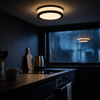 Round black flush mount ceiling light lit over a kitchen worktop with rain on the dark window