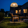 Tall woven solar garden floor lantern lit on a lawn beside patio chairs at blue hour